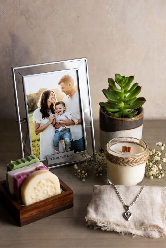 Elegant arrangement of sentimental gifts featuring a silver photo frame with family picture, handwritten "To Mom, With Love" tag, soy candle, small succulent plant, artisanal soap bars, and delicate heart necklace on neutral linen background.