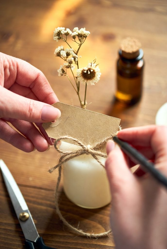 Close-up of a pair of hands personalizing a budget sympathy gift on a wooden table. One hand holds a small kraft paper gift tag while the other ties it with twine around a white candle jar decorated with dried flowers.